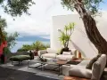 Arredare un terrazzo scoperto con divani chiari, tavolini geometrici e un pouf verde. Vista mare e scultura astratta.