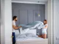 Coppia sorridente impegnata nel cambio lenzuola, con un piumone azzurro che svolazza sopra il letto.