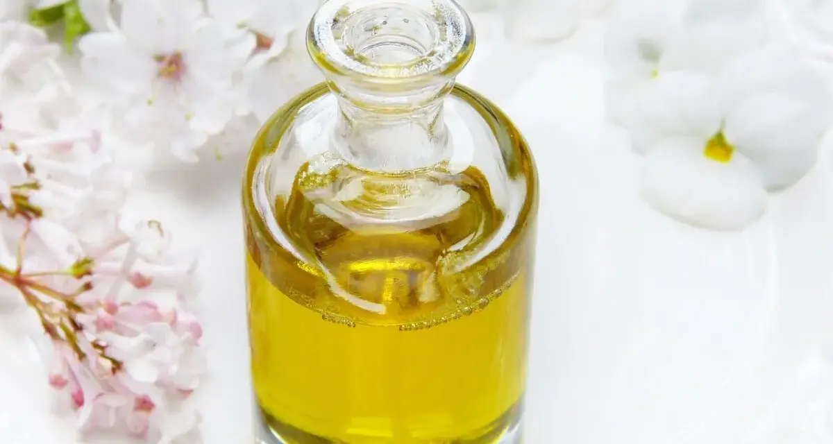 Flacone di vetro con liquido dorato, circondato da fiori bianchi e rosa. Un invito a pensare a dove buttare profumi pieni.