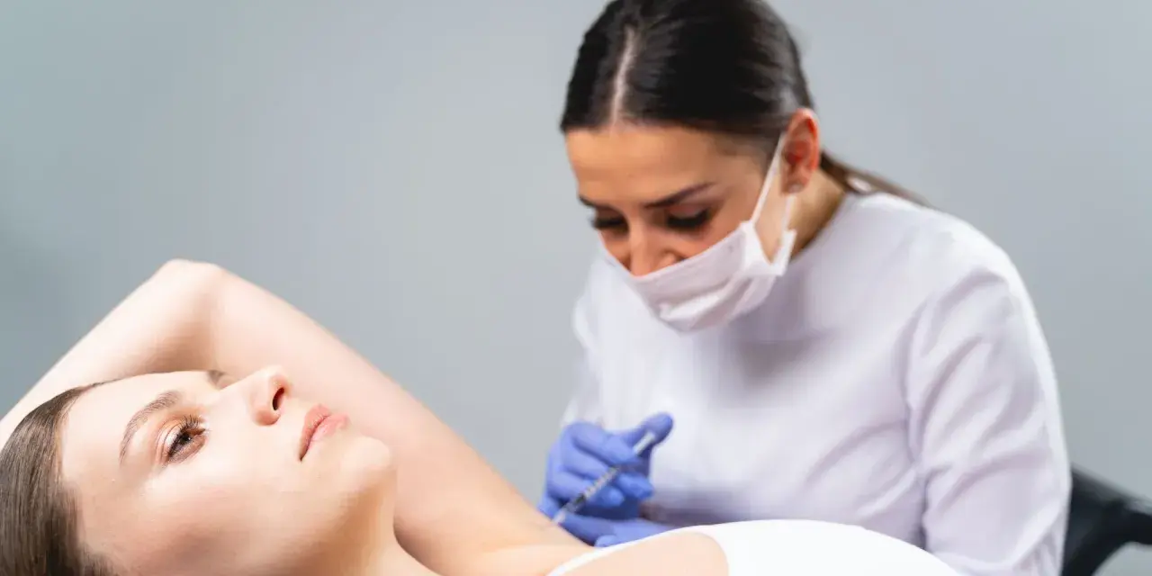 Una dottoressa con mascherina esegue un'iniezione all'ascella di una paziente. Un trattamento moderno, lontano dai sudore ascelle rimedi della nonna.