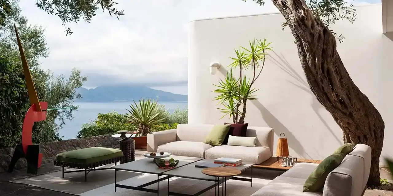 Arredare un terrazzo scoperto con divani chiari, tavolini geometrici e un pouf verde. Vista mare e scultura astratta.
