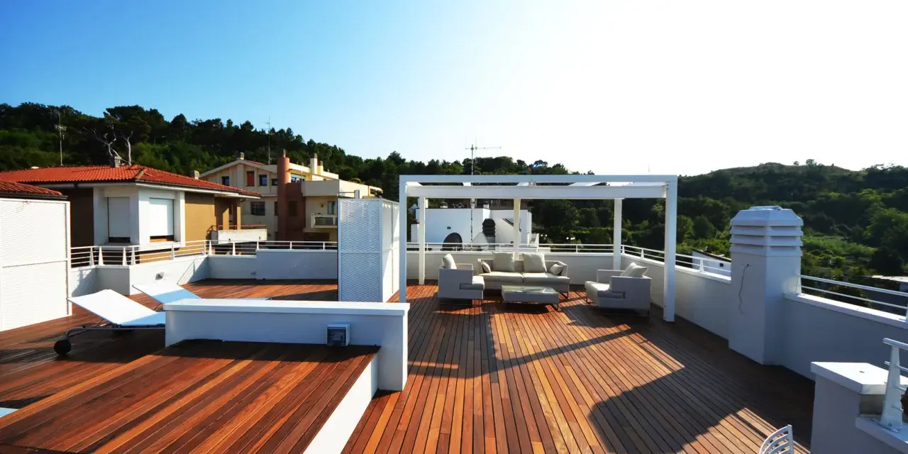 Terrazza sul tetto idee: un salotto all'aperto con divani, poltrone e tavolino sotto un pergolato bianco. Sdraio e vista su case e colline verdi.