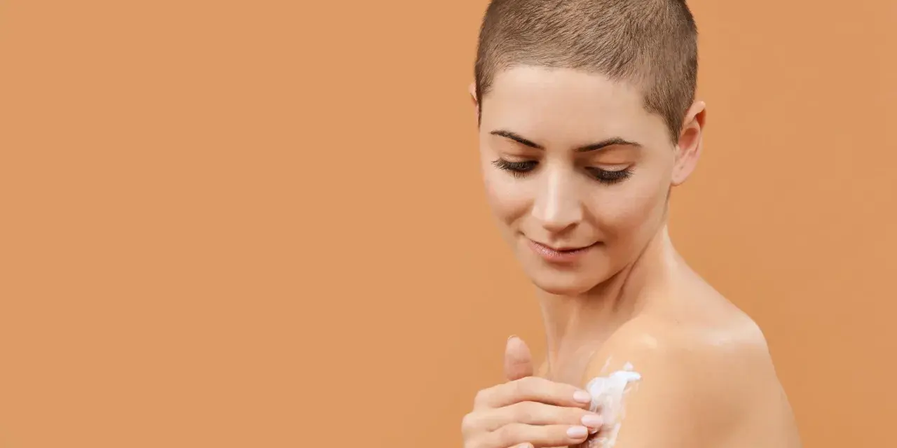 Donna con capelli corti applica crema idratante al burro di karitè viso sulla spalla, per una pelle morbida e nutrita.
