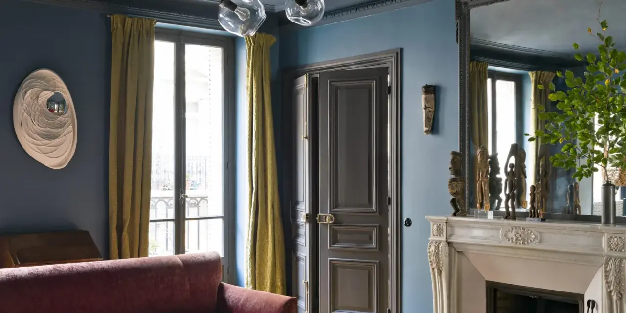 Soggiorno elegante con divano bordeaux, pareti blu balena e tendaggi dorati. Un camino in marmo e sculture africane completano l'arredo.