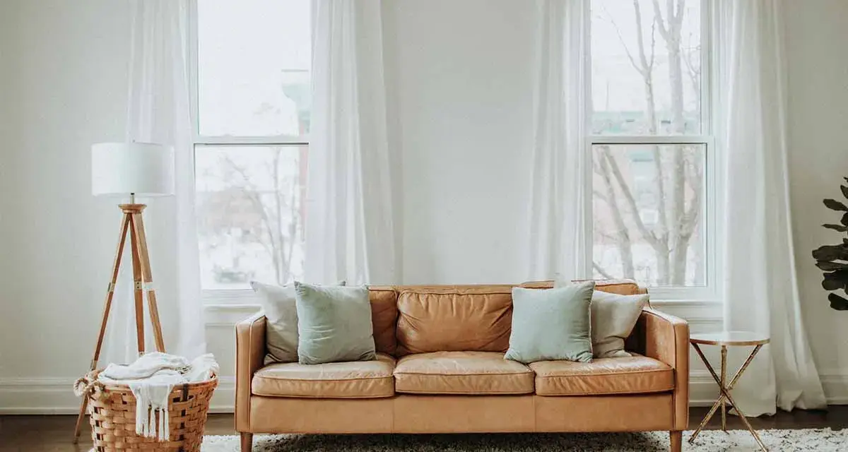 Un comodo divano in pelle color caramello è posizionato davanti alla porta finestra, con cuscini azzurri e un cesto di vimini con una coperta accanto.