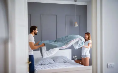 Coppia sorridente impegnata nel cambio lenzuola, con un piumone azzurro che svolazza sopra il letto.