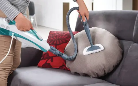 Persona usa un pulitore a vapore per pulire un cuscino del divano in tessuto. Scopri come pulire divano in tessuto con questo metodo efficace.