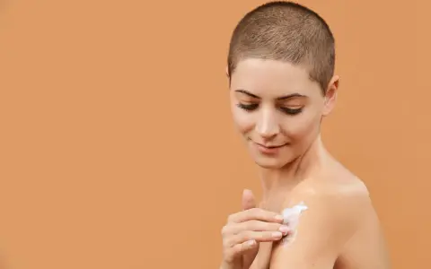 Donna con capelli corti applica crema idratante al burro di karitè viso sulla spalla, per una pelle morbida e nutrita.