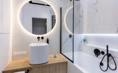 Moderno bagno con lavabo bianco su mobile in legno, specchio rotondo illuminato e vasca da bagno. Ottime bagno cieco idee.