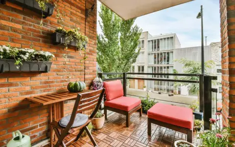 Arredare terrazzo appartamento: un angolo relax con sedie rosse, tavolino in legno e piante su muro di mattoni.