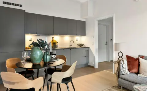 Soggiorno piccolo con divano e tavolo rotondo apparecchiato. Cucina moderna grigia con pensili illuminati.