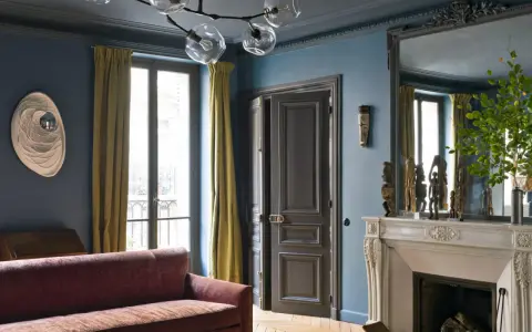Soggiorno elegante con divano bordeaux, pareti blu balena e tendaggi dorati. Un camino in marmo e sculture africane completano l'arredo.