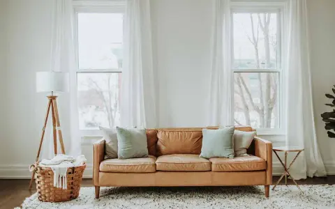 Un comodo divano in pelle color caramello è posizionato davanti alla porta finestra, con cuscini azzurri e un cesto di vimini con una coperta accanto.