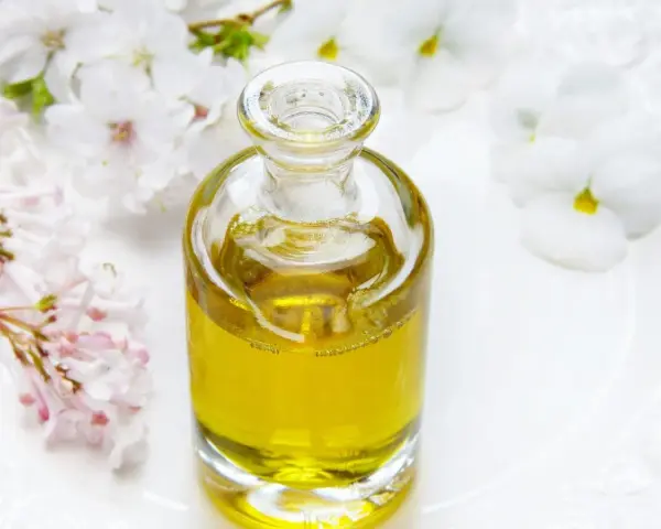 Flacone di vetro con liquido dorato, circondato da fiori bianchi e rosa. Un invito a pensare a dove buttare profumi pieni.