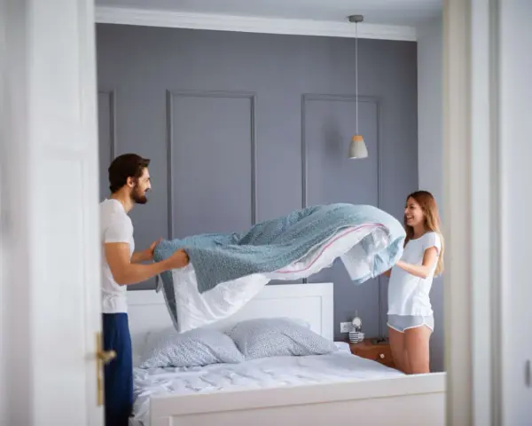 Coppia sorridente impegnata nel cambio lenzuola, con un piumone azzurro che svolazza sopra il letto.