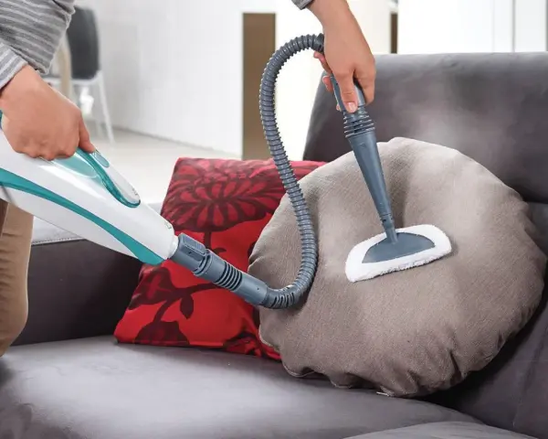 Persona usa un pulitore a vapore per pulire un cuscino del divano in tessuto. Scopri come pulire divano in tessuto con questo metodo efficace.