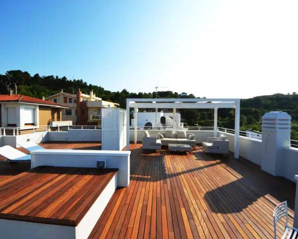 Terrazza sul tetto idee: un salotto all'aperto con divani, poltrone e tavolino sotto un pergolato bianco. Sdraio e vista su case e colline verdi.