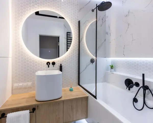 Moderno bagno con lavabo bianco su mobile in legno, specchio rotondo illuminato e vasca da bagno. Ottime bagno cieco idee.