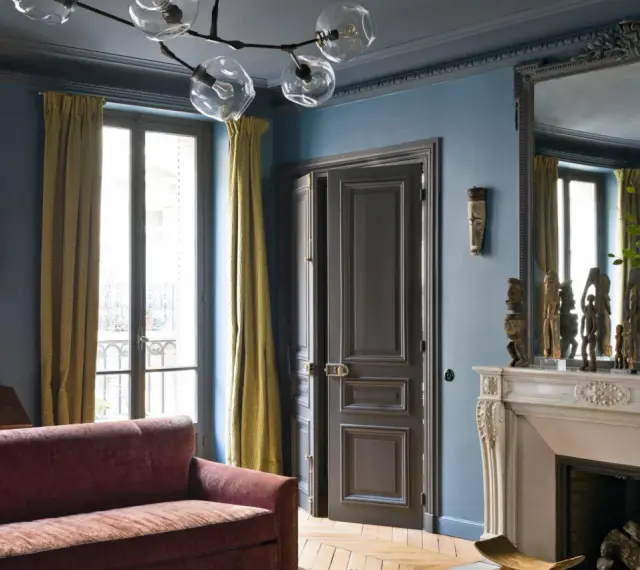 Soggiorno elegante con divano bordeaux, pareti blu balena e tendaggi dorati. Un camino in marmo e sculture africane completano l'arredo.