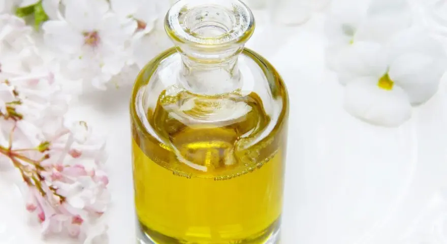 Flacone di vetro con liquido dorato, circondato da fiori bianchi e rosa. Un invito a pensare a dove buttare profumi pieni.