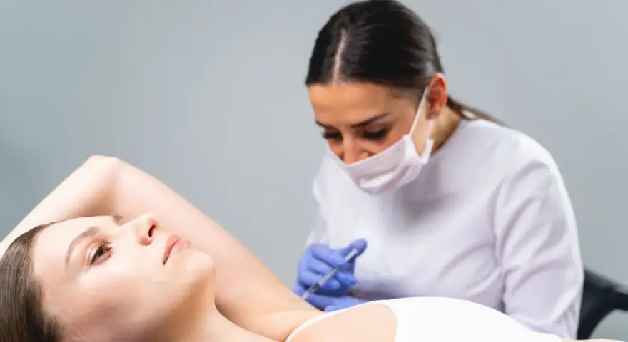 Una dottoressa con mascherina esegue un'iniezione all'ascella di una paziente. Un trattamento moderno, lontano dai sudore ascelle rimedi della nonna.