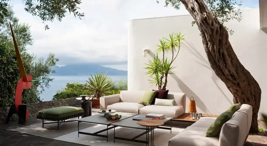 Arredare un terrazzo scoperto con divani chiari, tavolini geometrici e un pouf verde. Vista mare e scultura astratta.