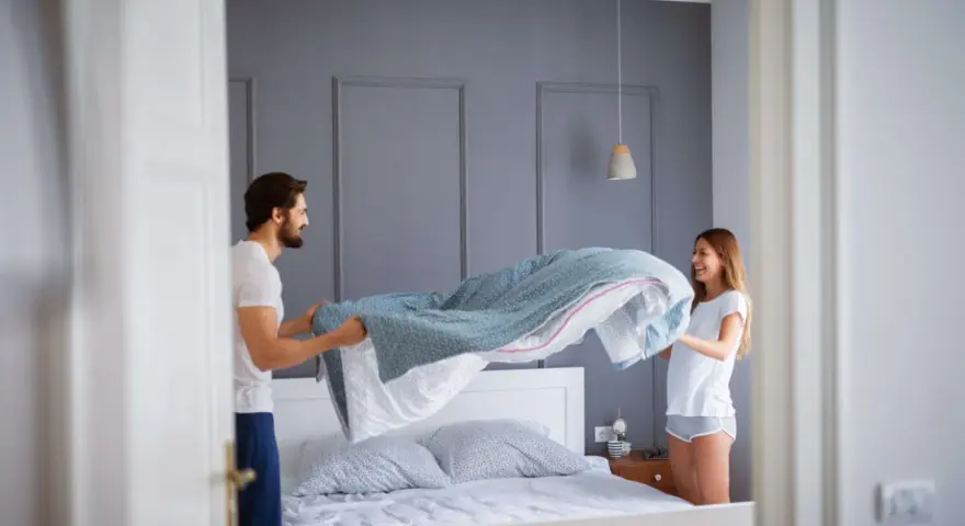 Coppia sorridente impegnata nel cambio lenzuola, con un piumone azzurro che svolazza sopra il letto.