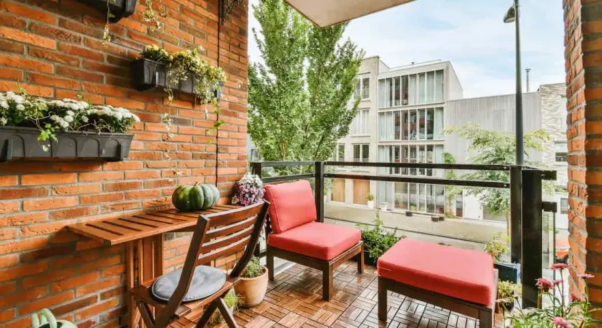 Arredare terrazzo appartamento: un angolo relax con sedie rosse, tavolino in legno e piante su muro di mattoni.