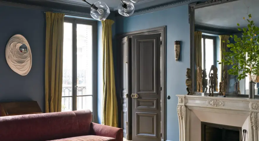 Soggiorno elegante con divano bordeaux, pareti blu balena e tendaggi dorati. Un camino in marmo e sculture africane completano l'arredo.