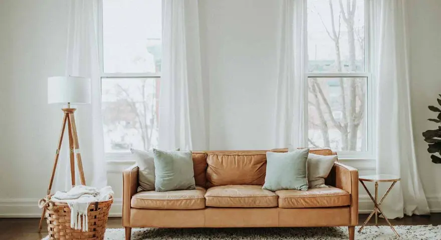 Un comodo divano in pelle color caramello è posizionato davanti alla porta finestra, con cuscini azzurri e un cesto di vimini con una coperta accanto.