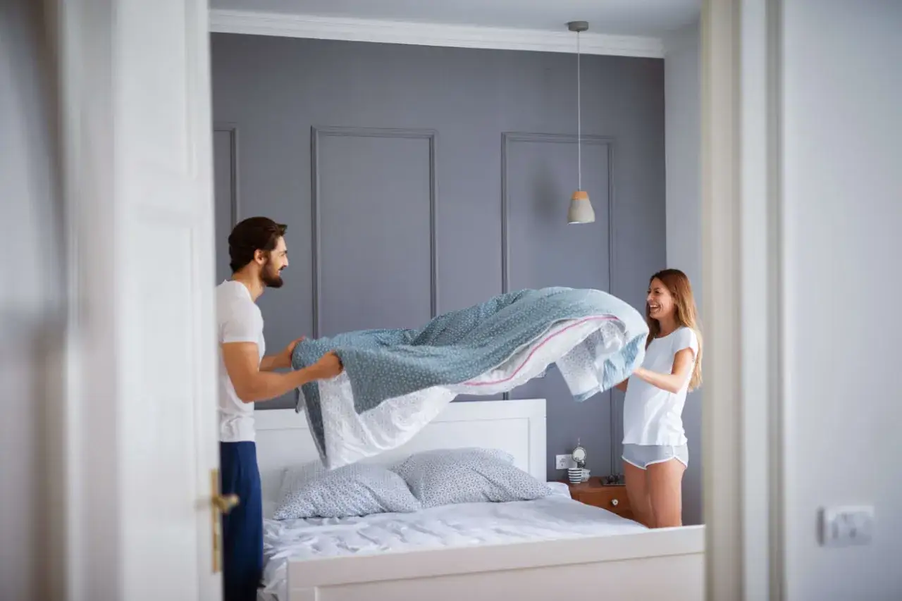 Coppia sorridente impegnata nel cambio lenzuola, con un piumone azzurro che svolazza sopra il letto.
