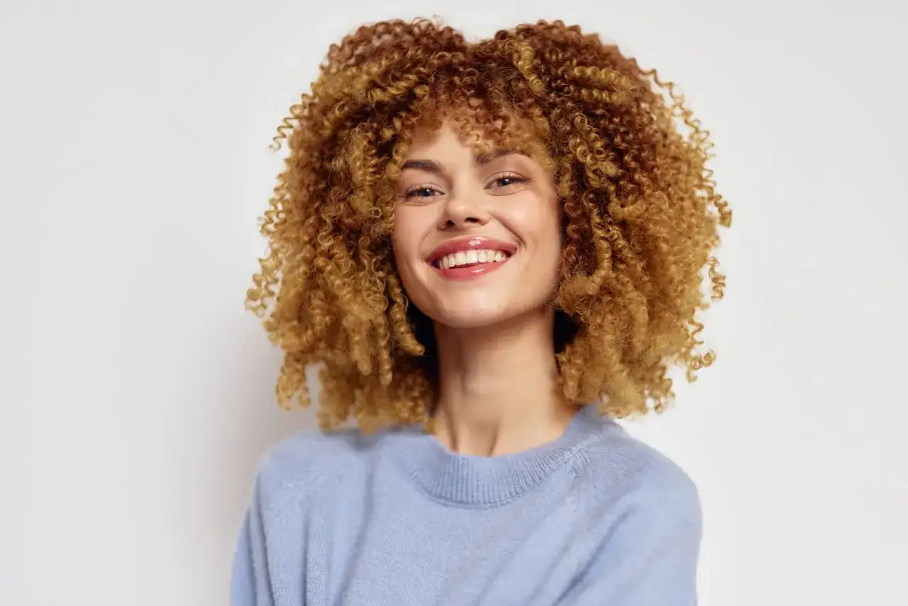 Sorriso radioso di una giovane donna con splendidi capelli mosci biondo ramati, indossa un maglione azzurro.