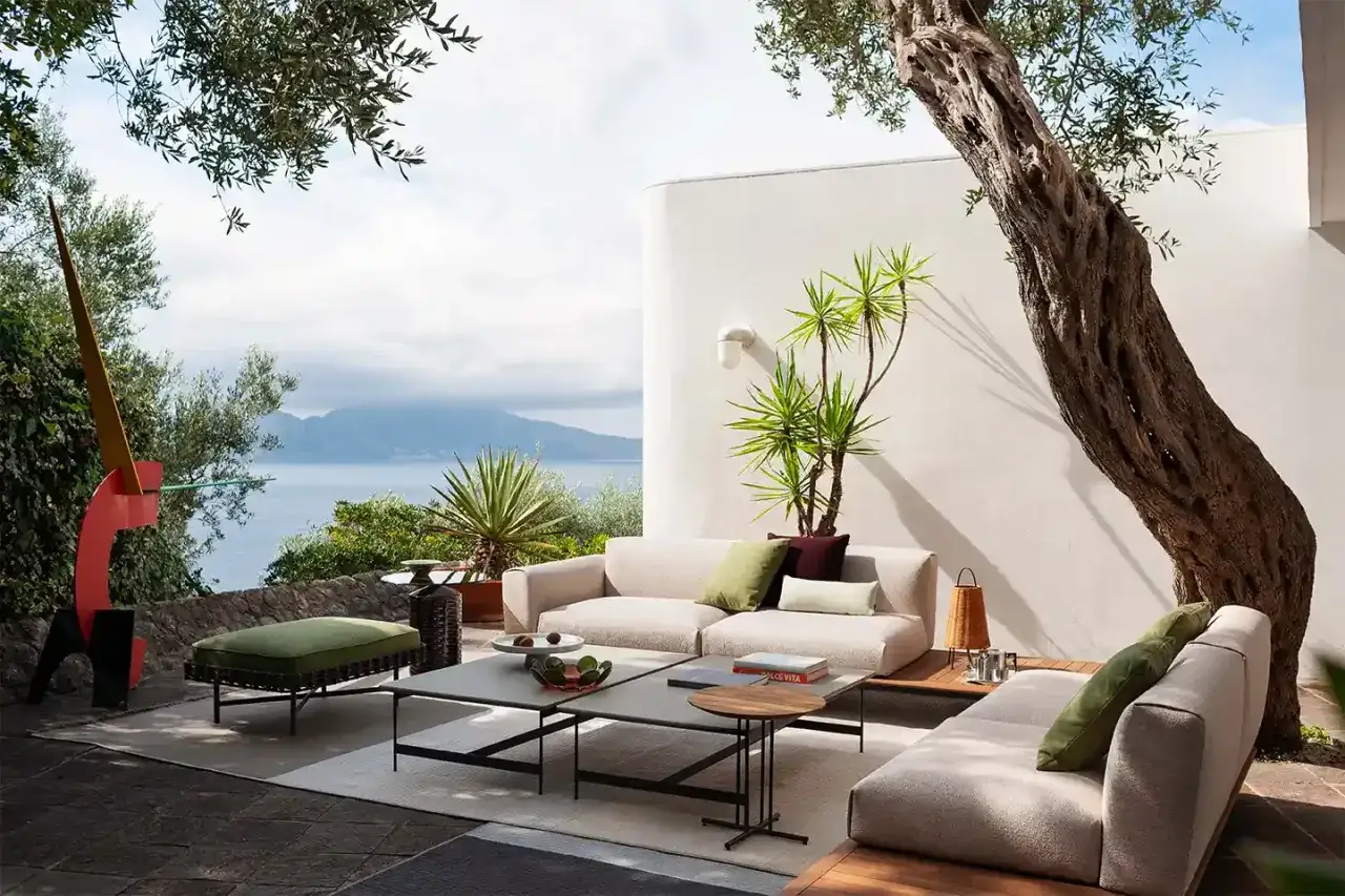 Arredare un terrazzo scoperto con divani chiari, tavolini grigi e un pouf verde. Vista mare e scultura colorata.