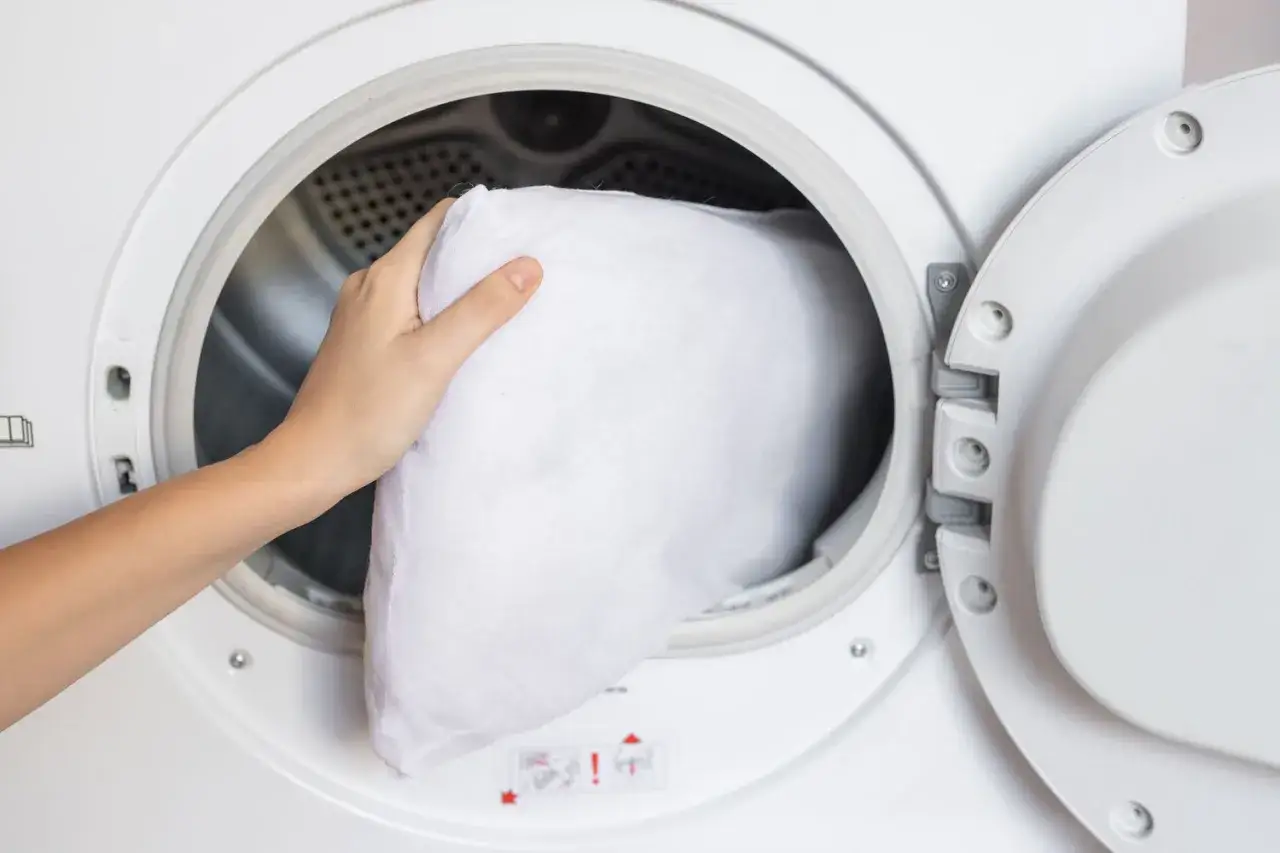 Mano inserisce un cuscino bianco nella lavatrice. Scopri come lavare i cuscini in lavatrice per mantenerli freschi e puliti.