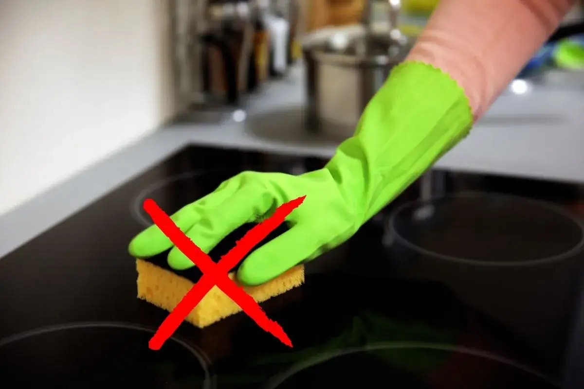 Mano con guanto verde che usa una spugna gialla sul piano cottura a induzione, con una grande X rossa sopra. Evita questo metodo per come pulire piano induzione.