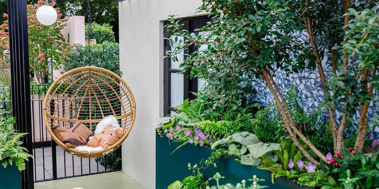 Un dondolo in rattan e cuscini invitano al relax. Un'oasi verde perfetta per arredare terrazzo appartamento, con piante rigogliose e fiori viola.